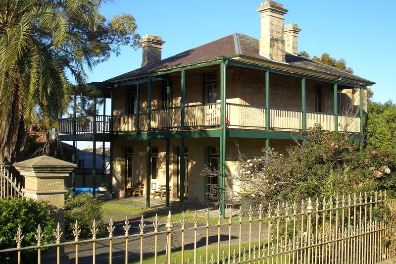 A two-storey sandstone house with wide verandahs behind an iron picket fence at Beverley Park