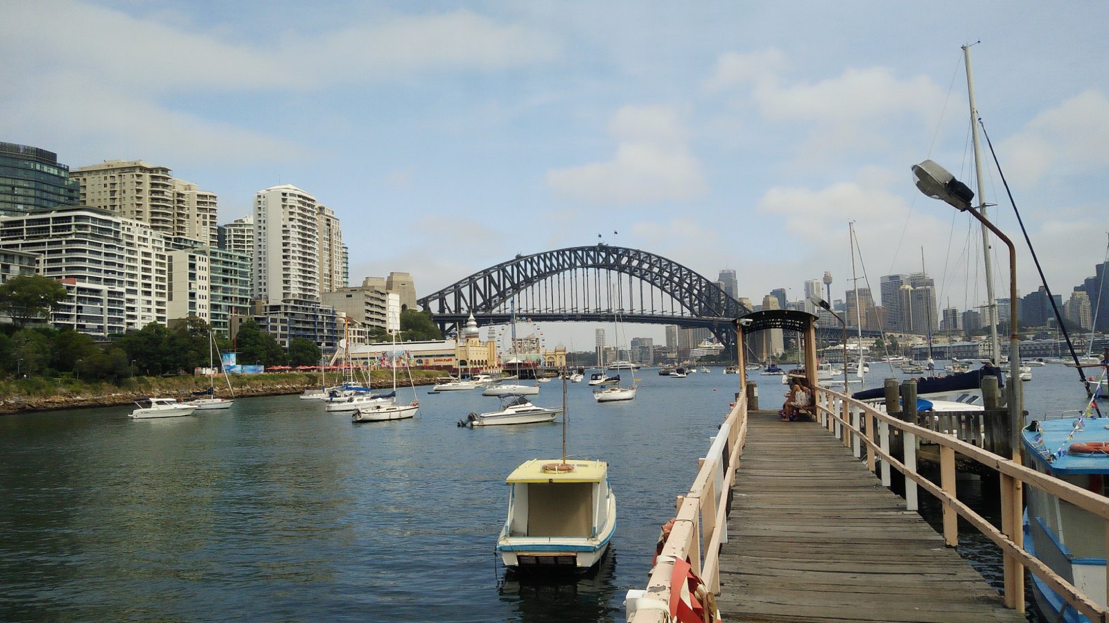 Timber jetty and moored boats in Lavender Bay, with Luna Park and the Sydney Harbour Bridge beyond.