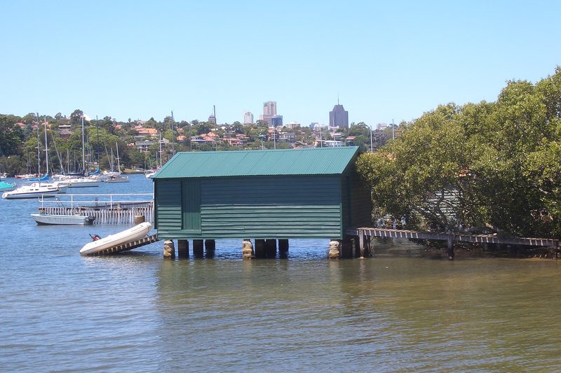 A green boatshed on Woodford Bay, Longueville, with the city skyline behind