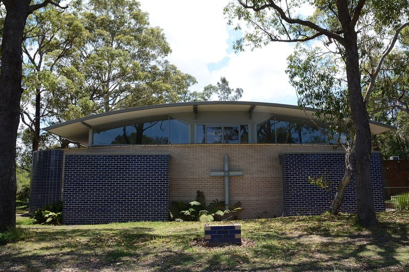 A mid-century church with a curved roof and cobalt brickwork, set among gums at South Turramurra